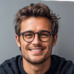 Smiling young man with glasses and tousled hair wearing a dark sweater against a gray background.